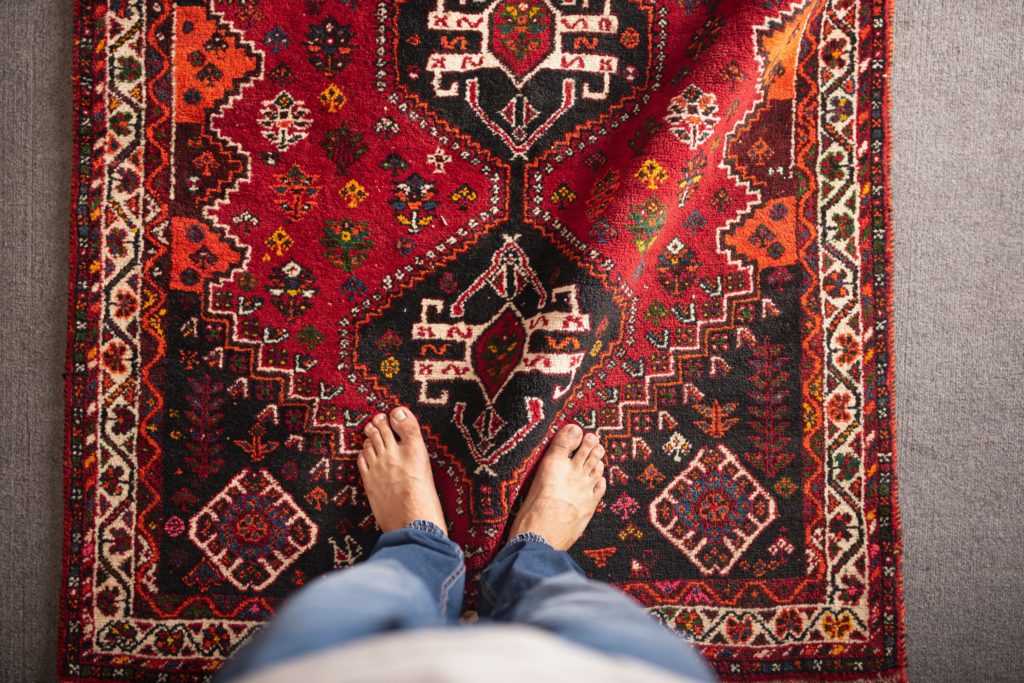 Bunched up persian rug on top of a loop carpet with man's feet in blue jeans standing on top